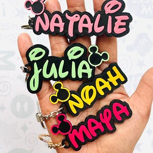 May include: Four colorful keychains with black Mickey Mouse ears and names "Natalie", "Julia", "Noah", and "Maya" written in a playful font.