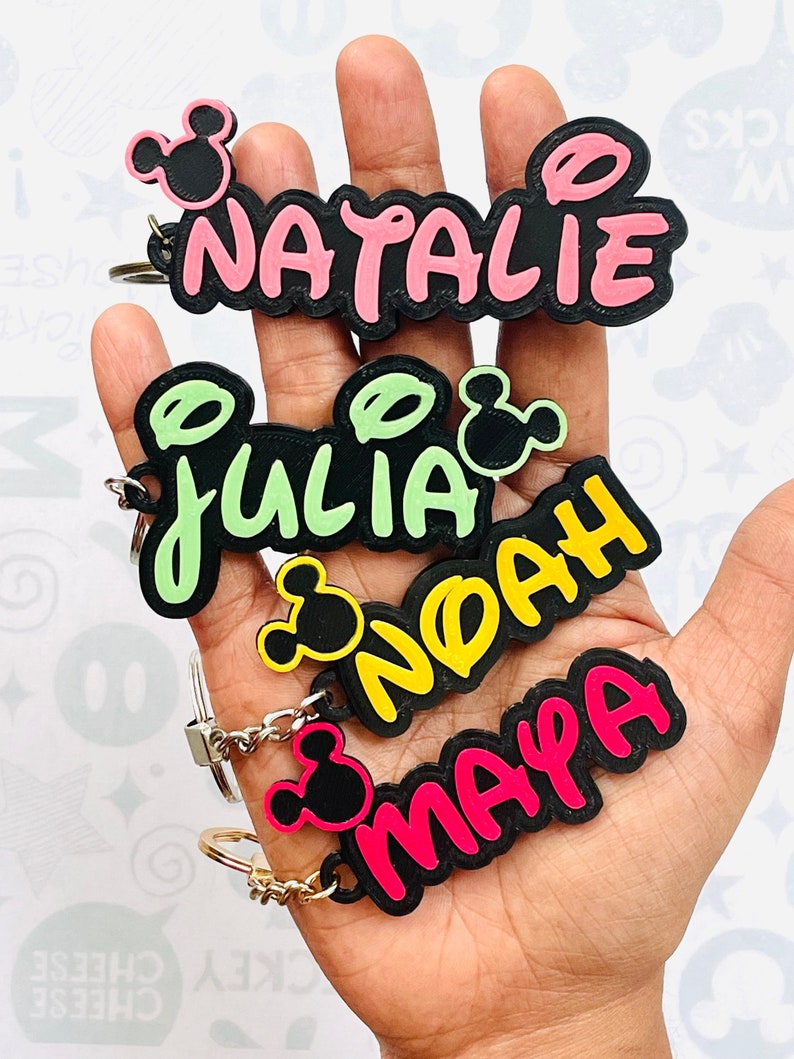 May include: Four colorful keychains with black Mickey Mouse ears and names "Natalie", "Julia", "Noah", and "Maya" written in a playful font.