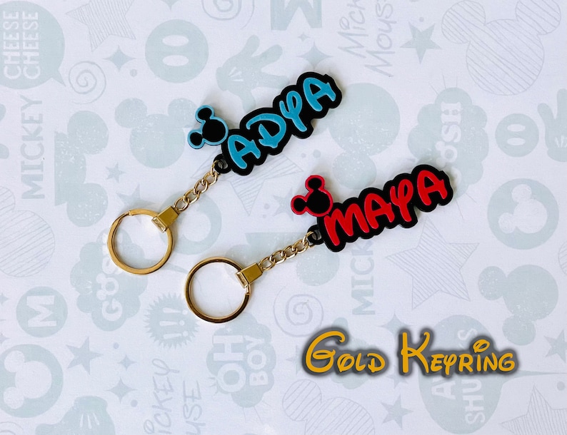 May include: Two gold keychains with black and blue Mickey Mouse ears. The keychains have the names "ADPA" in blue and "MAYA" in red.