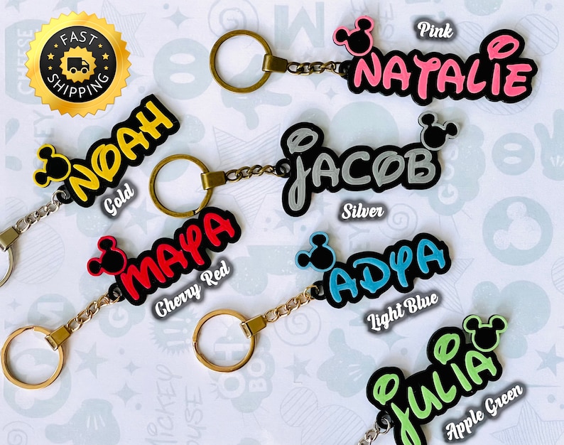 May include: A set of six personalized keychains with Mickey Mouse ears. The keychains are in different colors, including pink, silver, cherry red, light blue, apple green, and gold. Each keychain has a name on it: Natalie, Jacob, Maya, Adya, Julia, and Noah.
