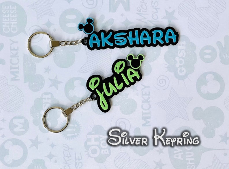 May include: Two silver keychains with black and green name tags. The name tags are shaped like a Mickey Mouse head and have the names "AKSHARA" and "JULIA" written in black letters. The keychains are on a white background with a Mickey Mouse pattern.