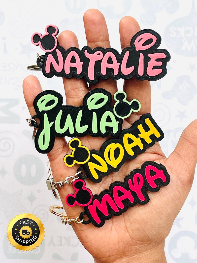 May include: Four personalized keychains with Mickey Mouse ears. The names Natalie, Julia, Noah, and Maya are written in black with pink, green, yellow, and pink outlines.