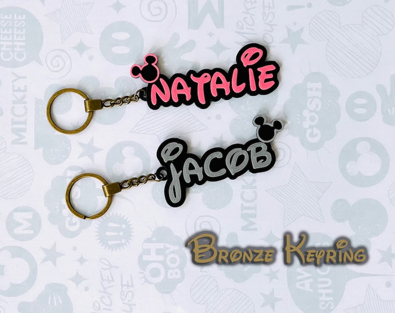 May include: Two bronze keychains with Mickey Mouse ears. One keychain has the name "Natalie" in pink letters and the other keychain has the name "Jacob" in gray letters. The keychains are on a white background with a Mickey Mouse pattern.