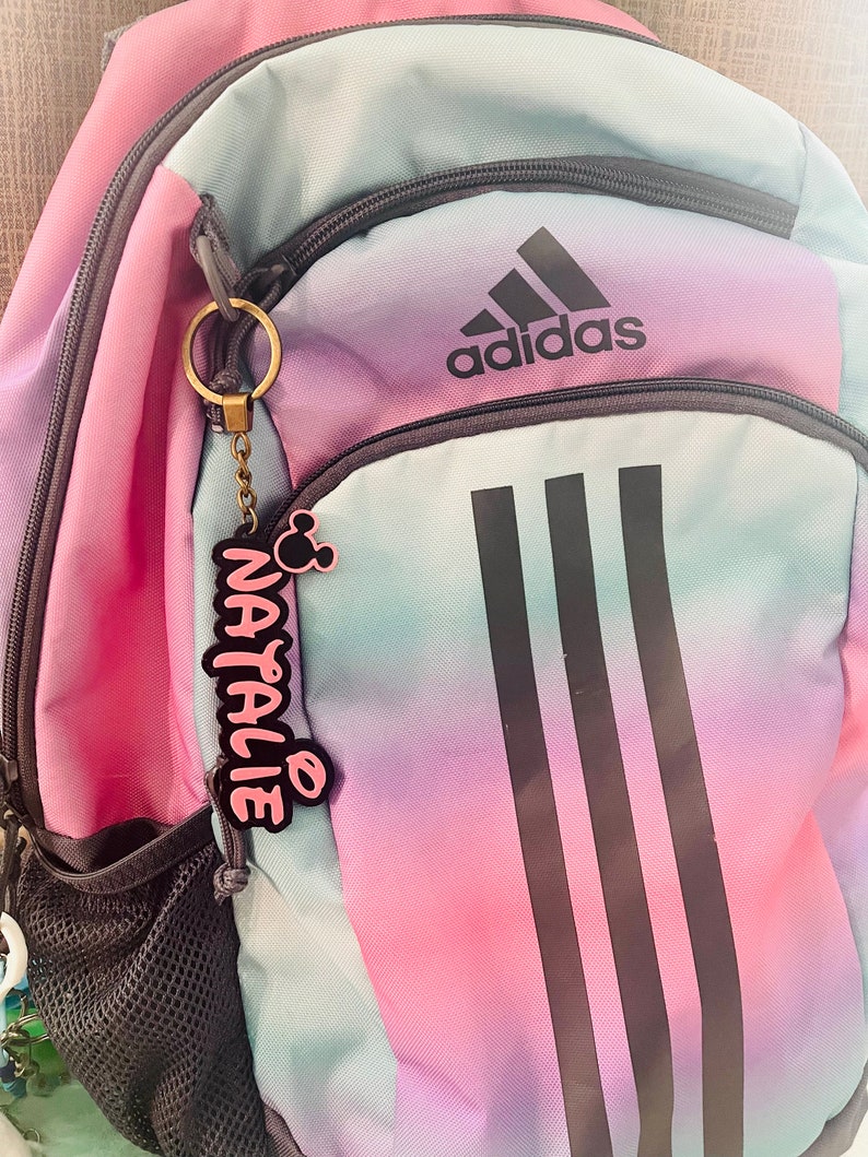 May include: A pink and blue ombre backpack with black stripes and the Adidas logo. A pink and black keychain with the name "Natalie" is attached to the zipper.
