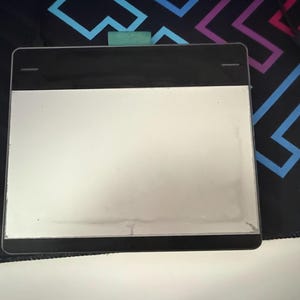 May include: A rectangular digital drawing tablet with a black top and a white drawing surface. A USB cable is visible in the background. The tablet is on a patterned surface.