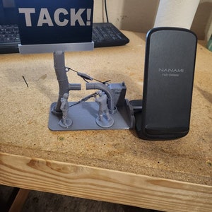 以下が含まれることがあります： 「TACK!」と書かれた看板と3体のミニチュアフィギュアが置かれたシーンの3Dプリントモデル。シーンは、黒いワイヤレス携帯電話充電器（「NANAMI FAST CHARGE」と表示）の隣にある木製のテーブルの上に置かれています。