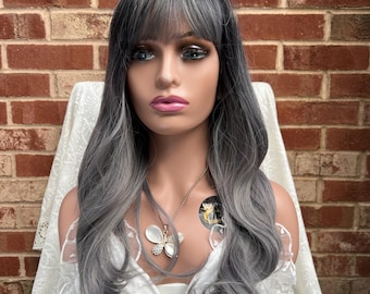 May include: A long, wavy, gray and silver wig with bangs. The wig is styled with loose waves and a side part. The wig is worn on a mannequin head.