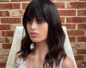 May include: A brown wavy bob wig with bangs. The wig is on a mannequin head and is styled with a side part. The wig is worn on a mannequin with a white lace top.