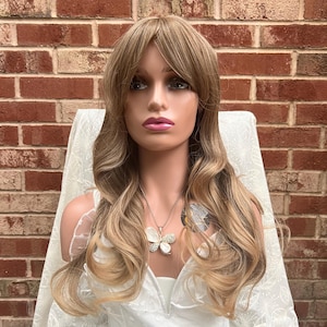 May include: A long, wavy, light brown wig with a fringe. The wig is styled in loose waves and has a natural-looking parting. The wig is worn by a mannequin with a white lace top and a silver butterfly necklace.