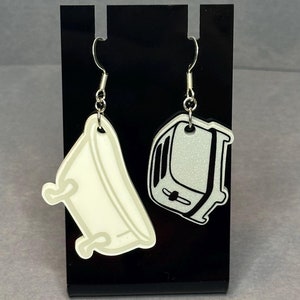 May include: A pair of dangle earrings featuring a white bathtub and a silver toaster. The earrings are hanging from a black display stand.