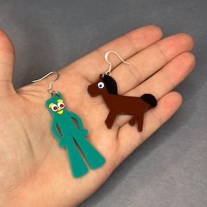 K&ouml;nnte beinhalten: Ein Paar Ohrringe mit einem gr&uuml;nen Comic-Charakter und einem braunen Pferd. Die Ohrringe sind aus Acryl und haben silberne Haken.