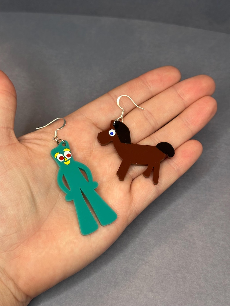 K&ouml;nnte beinhalten: Ein Paar Ohrringe mit einem gr&uuml;nen Comic-Charakter und einem braunen Pferd. Die Ohrringe sind aus Acryl und haben silberne Haken.