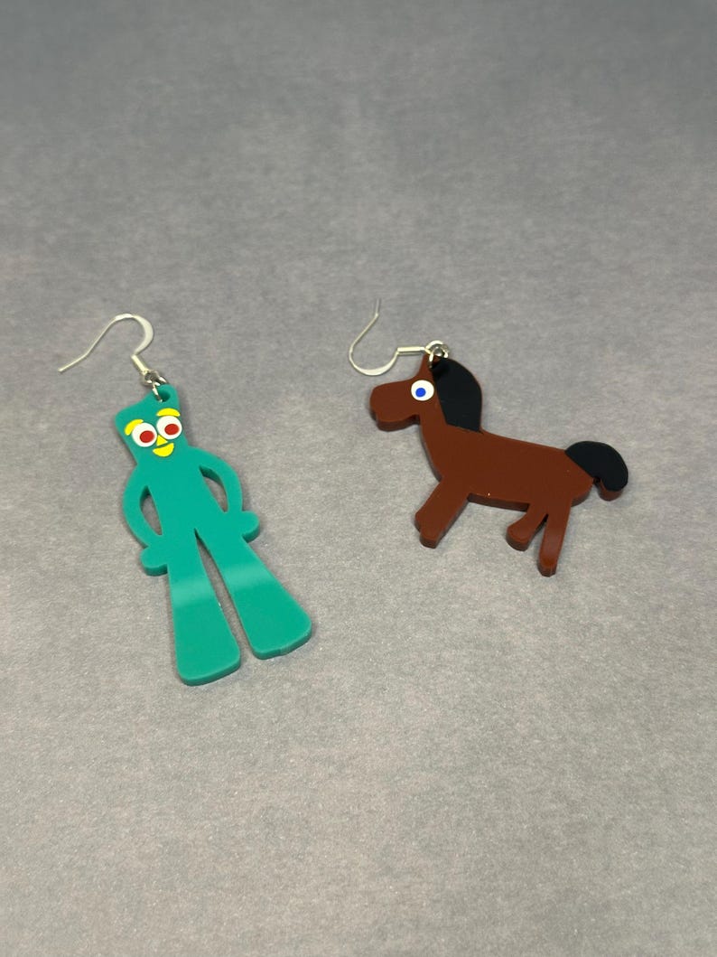 K&ouml;nnte beinhalten: Ein Paar Ohrringe mit einem gr&uuml;nen Comic-Charakter und einem braunen Pferd. Die Ohrringe sind aus Acryl und haben silberne Haken.