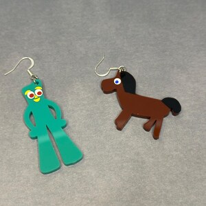 K&ouml;nnte beinhalten: Ein Paar Ohrringe mit einer gr&uuml;nen Strichm&auml;nnchenfigur mit roten Augen und einem braunen Pferd. Die Ohrringe sind aus Acryl und haben silberne Haken.
