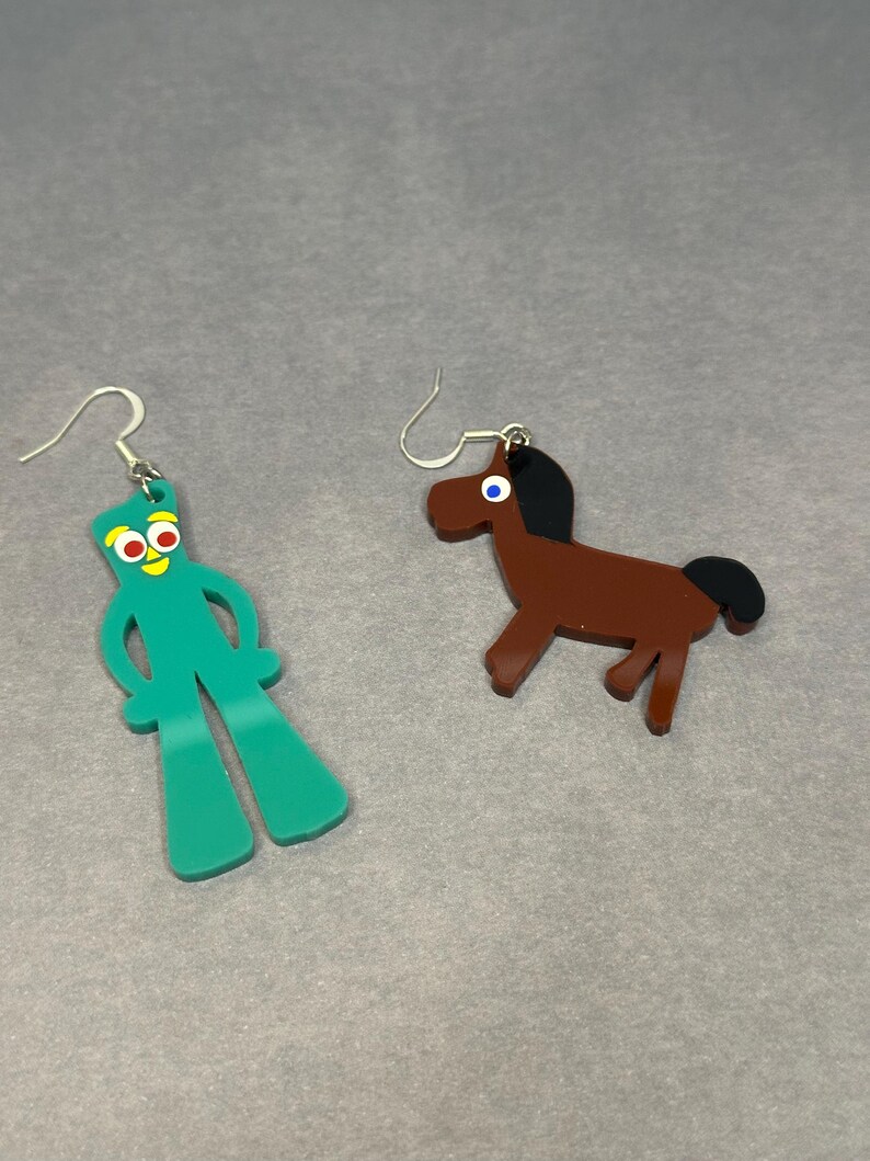 K&ouml;nnte beinhalten: Ein Paar Ohrringe mit einer gr&uuml;nen Strichm&auml;nnchenfigur mit roten Augen und einem braunen Pferd. Die Ohrringe sind aus Acryl und haben silberne Haken.