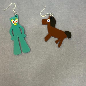 K&ouml;nnte beinhalten: Ein Paar H&auml;ngeohrringe. Ein Ohrring zeigt eine gr&uuml;ne Comicfigur mit gelben Augen und rotem Mund. Der andere Ohrring zeigt ein braunes Pferd mit schwarzer M&auml;hne und Schwanz.