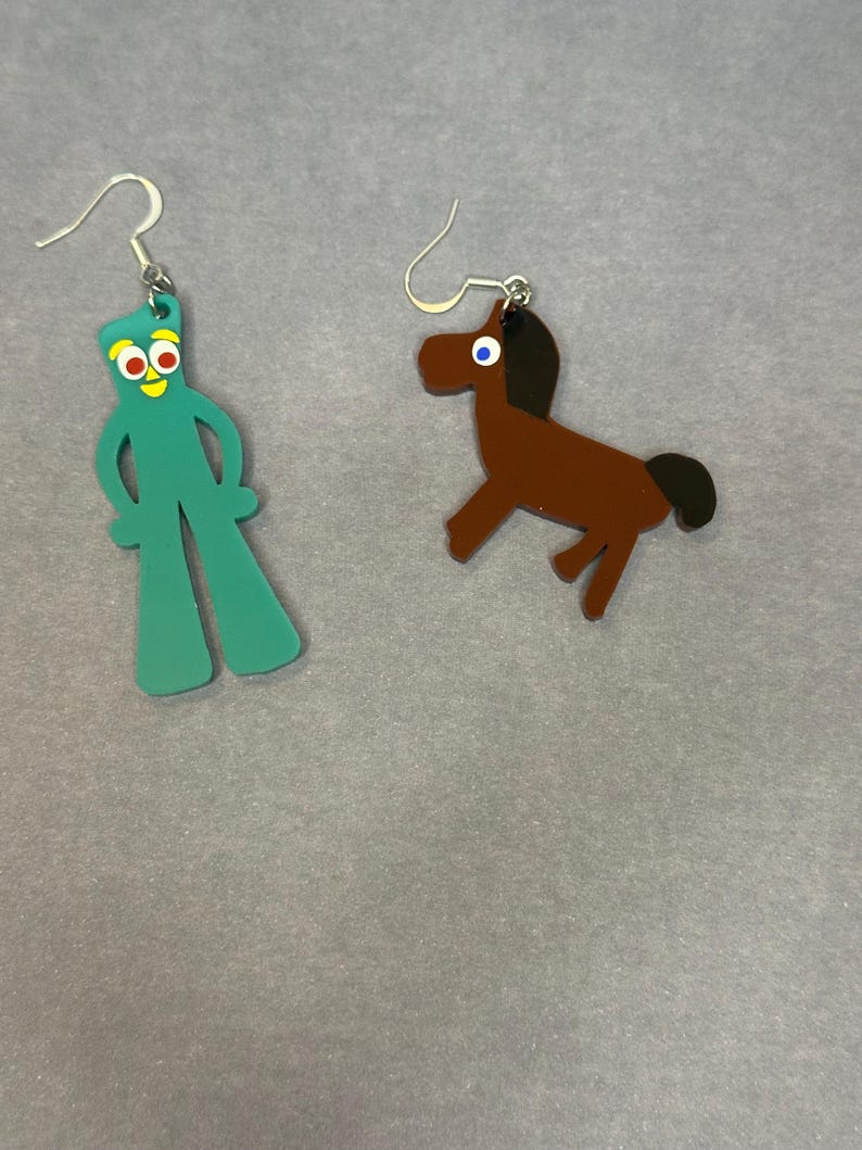 K&ouml;nnte beinhalten: Ein Paar H&auml;ngeohrringe. Ein Ohrring zeigt eine gr&uuml;ne Comicfigur mit gelben Augen und rotem Mund. Der andere Ohrring zeigt ein braunes Pferd mit schwarzer M&auml;hne und Schwanz.