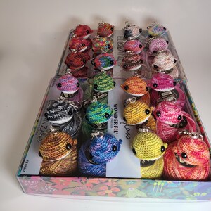 May include: A clear plastic box filled with colorful crocheted fish keychains. The keychains are made with different colors of yarn, including red, blue, green, yellow, and purple. The box is labeled with the words "Wonderfil" and "Eleganza".