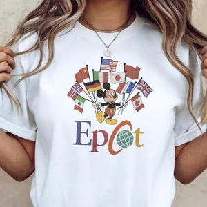 Op de afbeelding: Wit T-shirt met Mickey Mouse die vlaggen van verschillende landen vasthoudt. Het woord "Epcot" staat in kleurrijke letters eronder. Het T-shirt wordt gedragen met een blauwe spijkerbroek en witte sneakers.