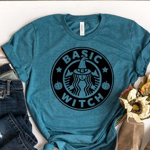 May include: A teal short-sleeved t-shirt featuring a black circular graphic with the text BASIC WITCH, a witch hat, and pumpkin icons. The shirt is displayed alongside dark blue distressed denim jeans and a pair of tan open-toe heeled shoes.