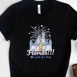 May include: Black t-shirt with a graphic of a fairytale castle, fireworks, and the text "Florida!!! no hell of a drug." Accessories include a watch, earrings, glasses, a plant, a coffee cup, and a brown purse.