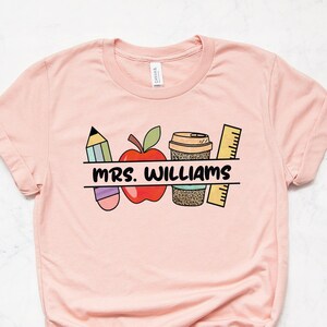 May include: A pink t-shirt with a graphic design of a pencil, apple, coffee cup, and ruler. The text "MRS. WILLIAMS" is printed on the shirt.