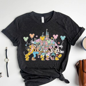 K&ouml;nnte beinhalten: Ein dunkelgraues T-Shirt mit einem bunten Motiv von Mickey Mouse, Minnie Mouse, Goofy und Pluto vor dem Schloss von Aschenputtel. Die Figuren halten Ballons und Sterne.
