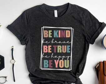 May include: Dark gray t-shirt with the words "BE KIND, be brave, BE TRUE, be happy, BE YOU" in a white rectangular frame. The shirt is styled with a watch, glasses, a plant, a cup of coffee, and a brown purse.