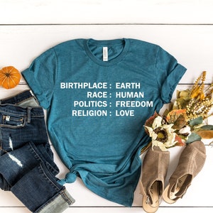 May include: A teal blue t-shirt with the text "Birthplace: Earth, Race: Human, Politics: Freedom, Religion: Love" printed in white on the front. The shirt is laid flat on a white wooden surface with a pair of blue jeans, a small orange pumpkin, and a bouquet of dried flowers.
