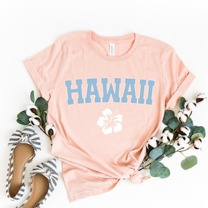 May include: A light pink t-shirt with the word "HAWAII" printed in blue and a white hibiscus flower graphic. The shirt is laid flat on a white background with a sprig of eucalyptus and cotton bolls.