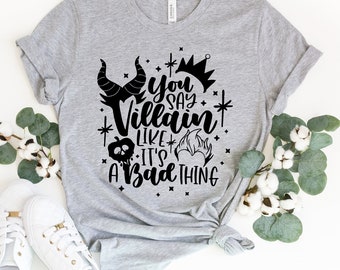 Puede incluir: Una camiseta gris jaspeado con el texto negro "You say Villain like it's a bad thing". El diseño incluye cuernos, una corona, una calabaza y una llama. Zapatillas blancas y ramitas verdes rodean la camiseta.
