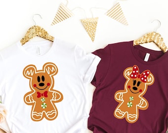 Puede incluir: Dos camisetas, una blanca y otra burdeos, cada una con un diseño de personaje de pan de jengibre. La camiseta blanca tiene un hombre de jengibre con pajarita roja. La camiseta burdeos tiene una mujer de jengibre con lazo rojo.