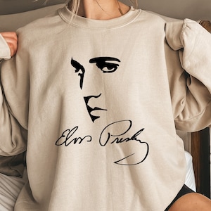 Pode incluir: Camisola bege com um desenho a tra&ccedil;o preto do rosto e da assinatura de Elvis Presley. O desenho inclui os seus olhos, nariz e boca, com o nome "Elvis Presley" escrito abaixo em it&aacute;lico. A camisola &eacute; de cor neutra.