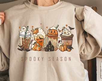 May include: Beige sweatshirt with a Halloween-themed graphic. The design features cartoon coffee drinks with pumpkin and ghost details, bats, and the words "Spooky Season" in brown letters. The sweatshirt has long sleeves.