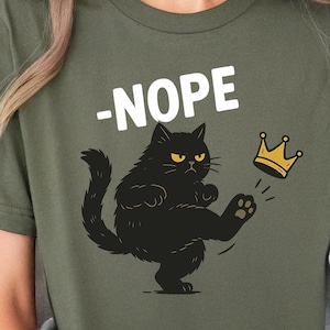 May include: Olive green t-shirt with a black cat kicking a gold crown. The word "-NOPE" is printed above the cat. The shirt is made of a soft material.