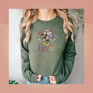 Op de afbeelding: Olijfgroene sweatshirt met een afbeelding van Mickey Mouse met vlaggen van over de hele wereld. Het woord "Epcot" staat eronder in kleurrijke letters. De sweatshirt heeft lange mouwen en een ronde hals.