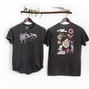 May include: Two dark grey t-shirts hanging on a wooden rod. The front of the first shirt features a cartoon design with the text "Everything you want to see." The back of the second shirt has a cartoon design.