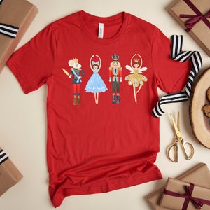 May include: A red t-shirt featuring a whimsical illustration of characters from The Nutcracker ballet. The design includes a mouse king, a ballerina, a nutcracker, and a fairy. The shirt is surrounded by wrapped gifts and festive decorations.