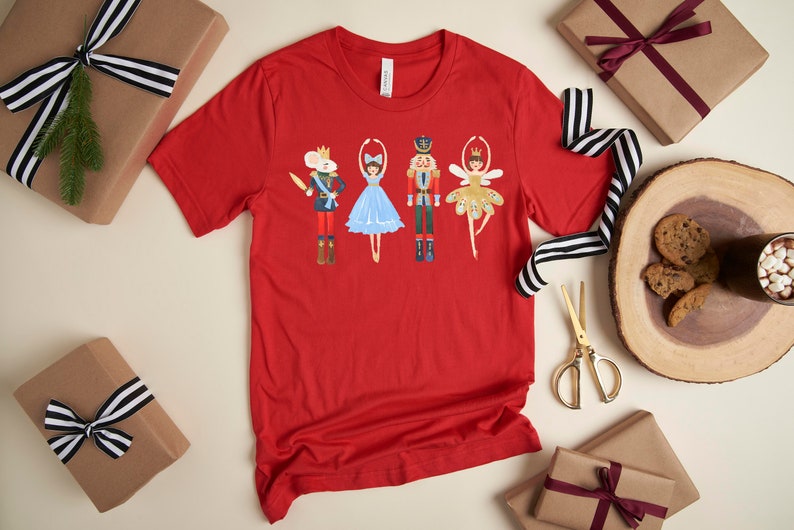 May include: A red t-shirt featuring a whimsical illustration of characters from The Nutcracker ballet. The design includes a mouse king, a ballerina, a nutcracker, and a fairy. The shirt is surrounded by wrapped gifts and festive decorations.