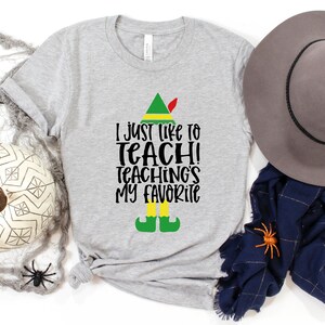 May include: A heather gray t-shirt with the text "I just like to teach! Teaching's my favorite" in black. The design includes an elf hat, shoes, and legs. A decorative hat, scarf, and spiders are also in the image.