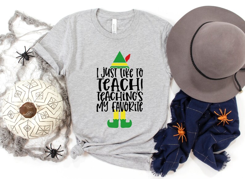 May include: A heather gray t-shirt with the text "I just like to teach! Teaching's my favorite" in black. The design includes an elf hat, shoes, and legs. A decorative hat, scarf, and spiders are also in the image.