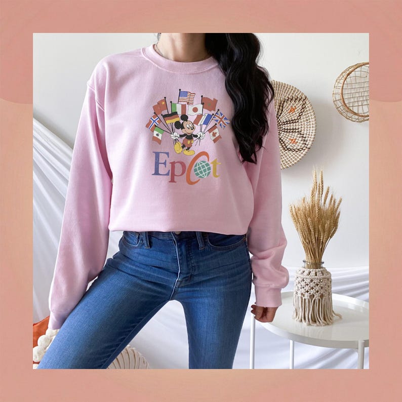 Op de afbeelding: Een lichtroze sweatshirt met een afbeelding van Mickey Mouse omringd door vlaggen en het woord "Epcot" in kleurrijke letters. Het sweatshirt wordt gecombineerd met een blauwe jeans.