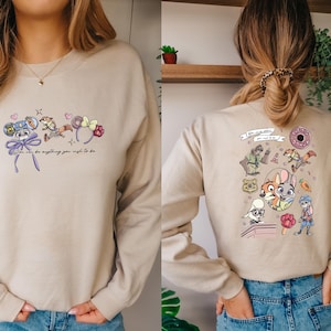 May include: Beige sweatshirt with cartoon graphics on the front and back. The front features mouse ears and the text "You can be anything you wish to be." The back has cartoon characters and the text "The Big Donut."
