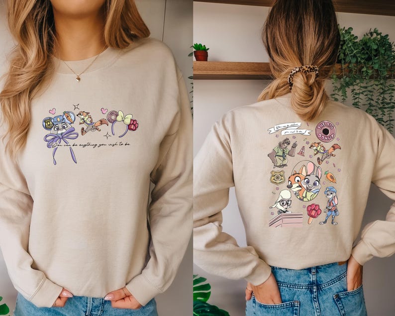 May include: Beige sweatshirt with cartoon graphics on the front and back. The front features mouse ears and the text "You can be anything you wish to be." The back has cartoon characters and the text "The Big Donut."