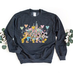 K&ouml;nnte beinhalten: Ein dunkelgraues Sweatshirt mit einem bunten Grafikdruck von Mickey Mouse, Minnie Mouse, Pluto, Goofy und Donald Duck vor dem Schloss von Aschenputtel. Die Figuren halten Ballons und Sterne sind im Hintergrund.