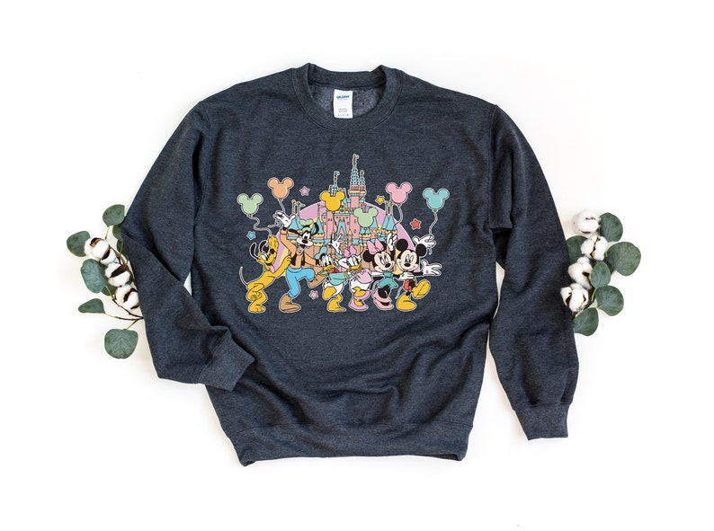 K&ouml;nnte beinhalten: Ein dunkelgraues Sweatshirt mit einem bunten Grafikdruck von Mickey Mouse, Minnie Mouse, Pluto, Goofy und Donald Duck vor dem Schloss von Aschenputtel. Die Figuren halten Ballons und Sterne sind im Hintergrund.