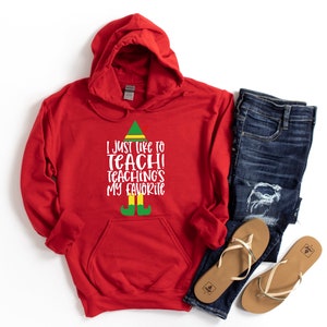 May include: A red hooded sweatshirt with the text "I just like to teach! Teaching's my favorite" in white, with an elf-themed design. The image also includes blue jeans and a pair of tan sandals.