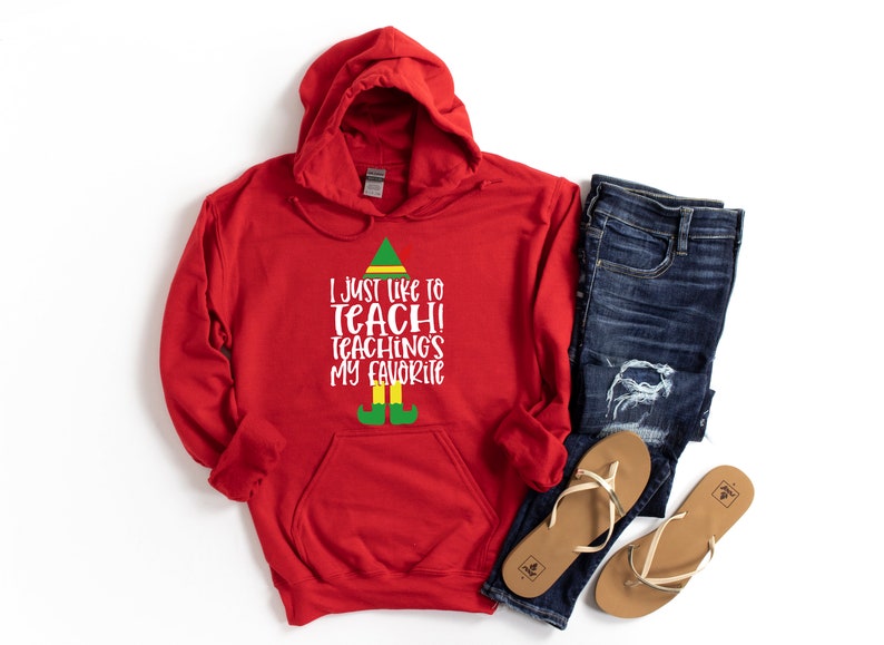 May include: A red hooded sweatshirt with the text "I just like to teach! Teaching's my favorite" in white, with an elf-themed design. The image also includes blue jeans and a pair of tan sandals.