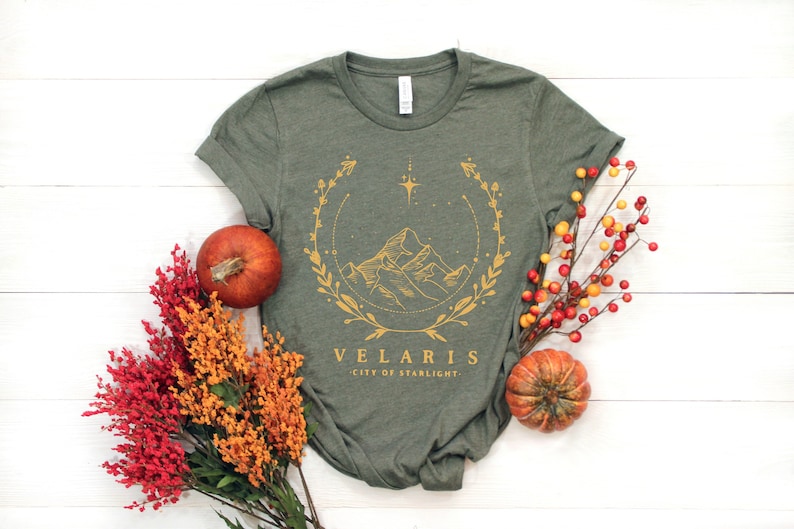 May include: A green t-shirt with a gold graphic of a mountain range, a star, and a wreath. The text "VELARIS CITY OF STARLIGHT" is printed below the graphic.