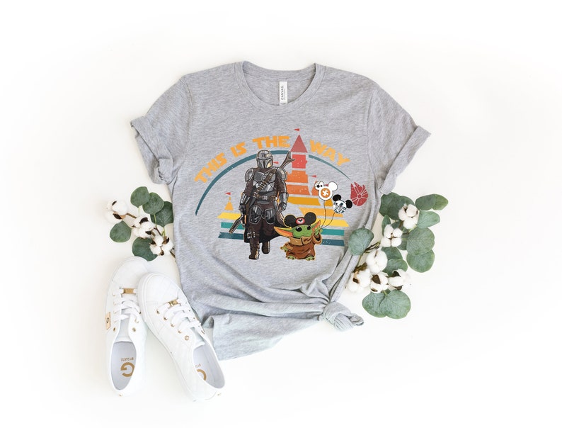K&ouml;nnte beinhalten: Ein graues T-Shirt mit einer Grafik von The Mandalorian und Grogu, mit dem Text "THIS IS THE WAY" in Orange. Das Design umfasst ein Schloss, Luftballons und einen farbenfrohen Sonnenuntergang. Wei&szlig;e Turnschuhe und Baumwollzweige sind ebenfalls im Bild.